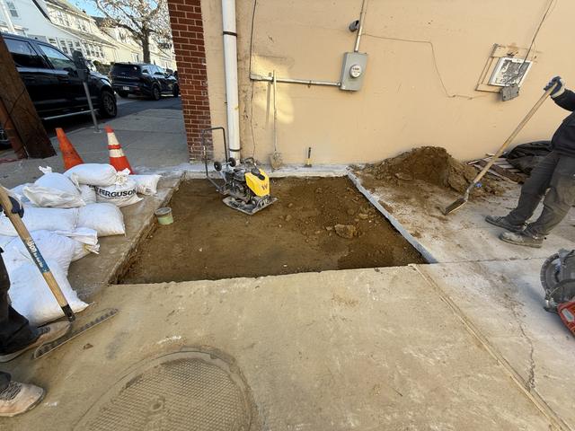 Excavated and compacted base beside a brick building, with a plate compactor, sandbags and safety cones